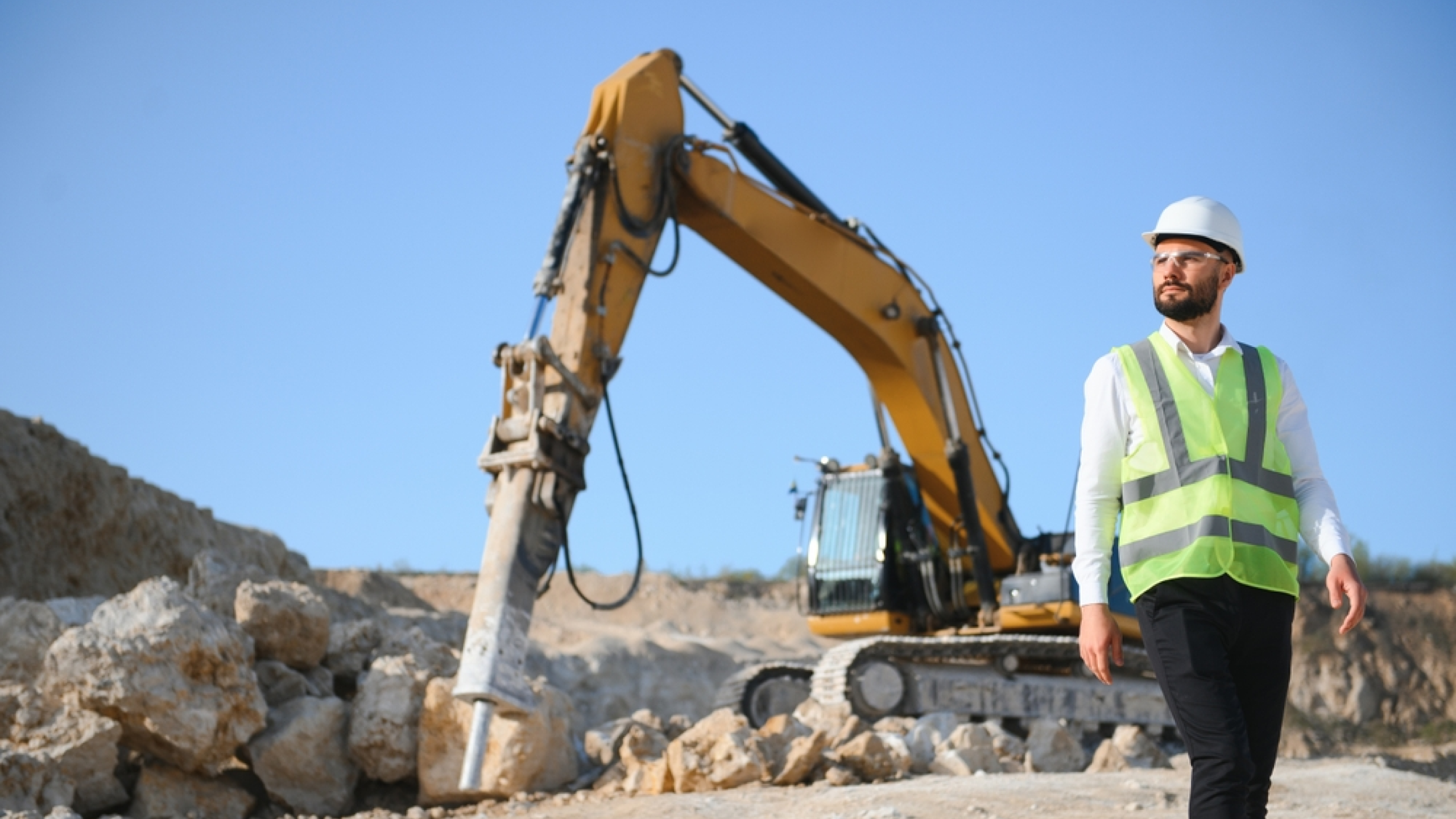 Excavator fitted with a hydraulic breaker working broken concrete while a worker in high-visibility clothing observes