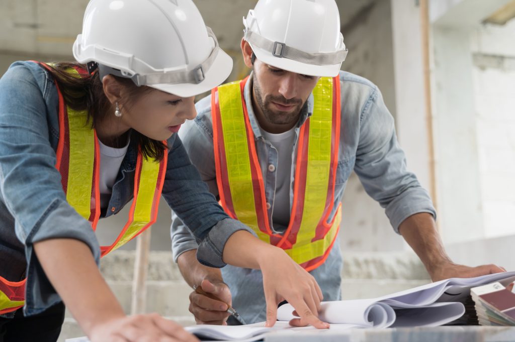 Two construction workers in hard hats and high-visibility vests examine blueprints on a construction site.