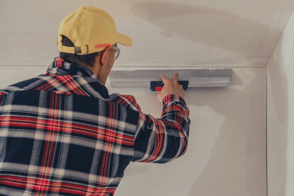 Construction worker in a yellow cap and plaid shirt smoothing plaster along a wall with a trowel-like tool while facing away from the camera.