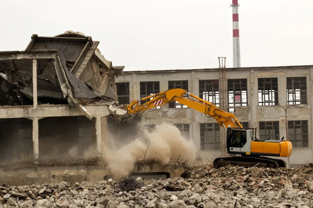 Excavator demolishing a concrete building, breaking walls and creating a cloud of dust amid scattered rubble.