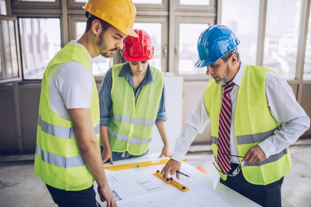 Three construction workers in high-visibility vests and hard hats study blueprints on a table in a bright indoor site.